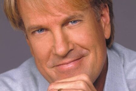 John Tesh smiles directly at the camera, hands clasped under his chin, wearing a gold wedding ring.