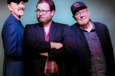 Three men in blazers and baseball caps pose against a gray and blue studio background, two smiling, one arms crossed.
