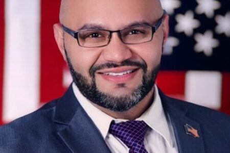 Rich Valdes. A Bald, bearded man in glasses, suit, and patterned tie smiles against an American flag backdrop.