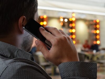 A man seen from behind holds a smartphone to his ear, talking on the phone, with a blurred background of warm lights.