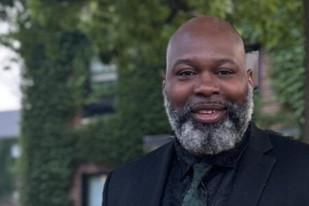 Smiling headshot of Ahmad Whitfield, a man with a salt-and-pepper beard, wearing a suit and tie, against a leafy background.