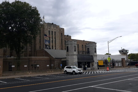 Exterior view of Auburn Correctional Facility with a street, parked cars, and a guard tower under a cloudy sky.