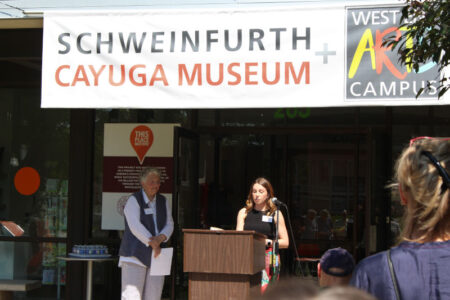 A woman speaks at a podium in front of a banner for Schweinfurth Art Center and Cayuga Museum, with another woman nearby.