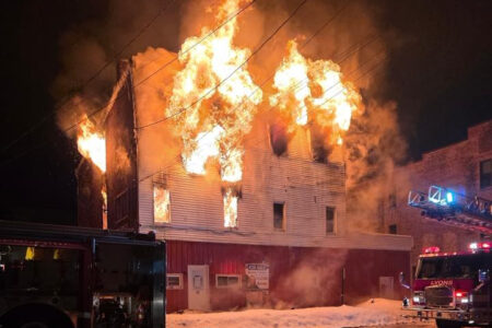A multi-story building in Lyons, NY, is engulfed in large flames at night, with snow on the ground and fire trucks nearby.