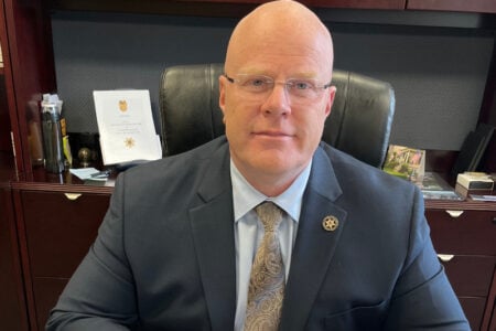 Wayne County Sheriff Robert Milby, a bald man in glasses, suit, and sheriff's badge, poses in his office.
