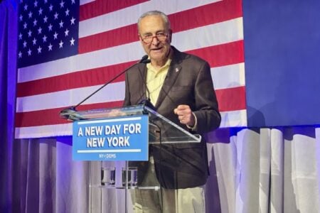 Senator Chuck Schumer speaks at a podium with an American flag and a 'A New Day for New York' sign.