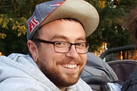 Smiling man with glasses, beard, and a New England Patriots hat, outdoors.