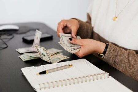 Person counting U.S. dollar bills at a desk with a planner and pen.