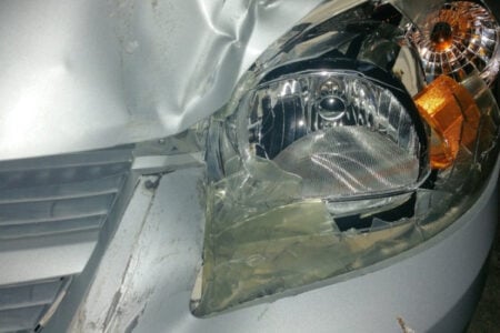 Close-up of a silver car's front end with a smashed headlight and dented body after a collision.