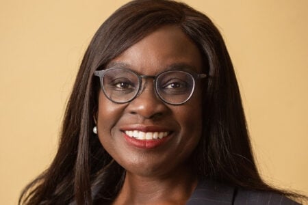 Headshot of a smiling Black woman with glasses and long dark hair, wearing a dark pinstripe suit against a plain background.