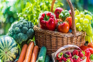 A wicker basket and other containers filled with fresh fruits and vegetables, including watermelon, carrots, and strawberries, outdoors.