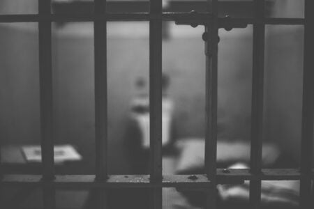 Close-up of jail cell bars in grayscale, with a blurry view of a bed and table inside a cell.