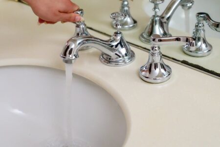 A hand turns on a chrome faucet, water flowing into a white sink on a beige countertop.