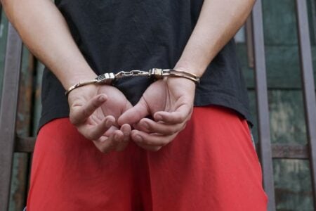 Person in handcuffs behind their back stands against a barred wall.