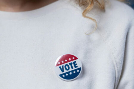 A person wears a patriotic 'VOTE' button with red, white, and blue stars and stripes on a white shirt.