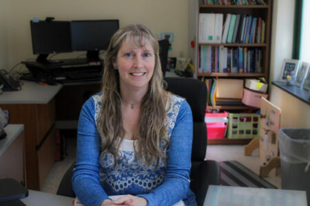 Anthea Teska, a woman with long blonde hair, smiles at the camera from her office desk with computers and a bookshelf.