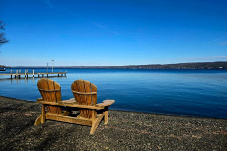 Two wooden Adirondack chairs on a pebbly shore face serene Canandaigua Lake under a clear blue sky, with a dock in the background.