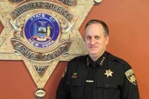 Cayuga County Sheriff Brian Schenck in uniform stands before the Sheriff's Office seal.