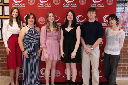 Six students, three female and three male, pose smiling in front of a red Cayuga Community College banner.