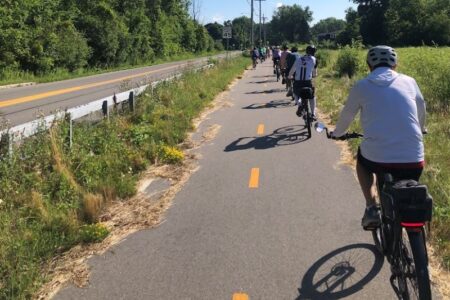 Cyclists ride single file on a paved trail with yellow lines, alongside a road and trees, under a sunny sky.