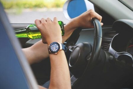 Person's hands on a steering wheel, one hand holding a green beer bottle to their mouth while driving.