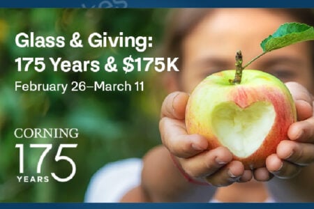 Hands hold an apple with a heart-shaped bite, promoting Corning's 'Glass & Giving' 175-year campaign.