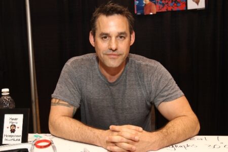 Actor Nicholas Brendon sits at a convention table, an autograph price list partially visible.