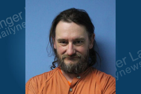 Mugshot of a man with long hair and a beard in an orange jumpsuit against a blue background.