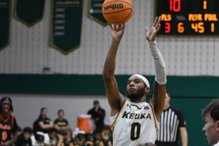 Keuka College basketball player #0, Lovelace, shoots a basketball during a game in a gymnasium with a scoreboard.