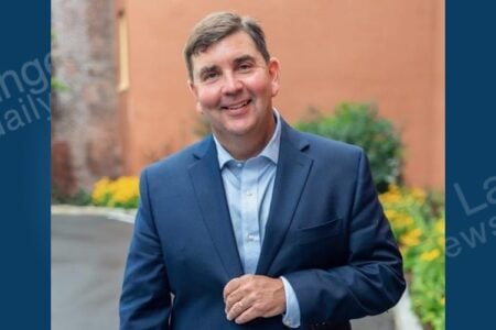 Headshot of Rep. Mannion, a smiling man in a blue suit, standing outdoors with brick buildings and greenery.