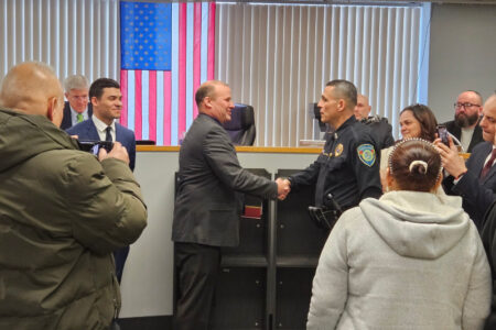 Agustin Gonzalez, in uniform, shakes hands with a man in a suit at his swearing-in ceremony, with an American flag behind them.