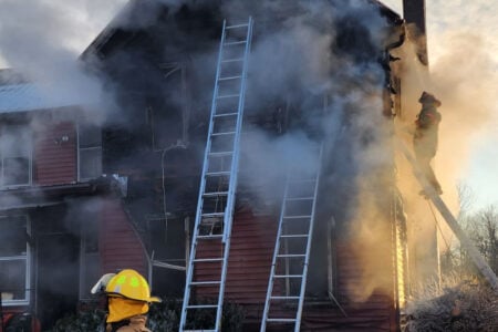 Firefighter watches as smoke and flames engulf a red house with two ladders propped against it.