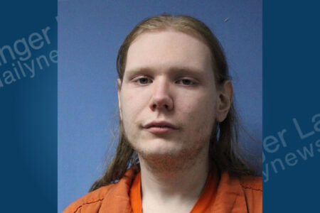 Mugshot of a man with long light hair and stubble, wearing an orange jail jumpsuit against a blue background.