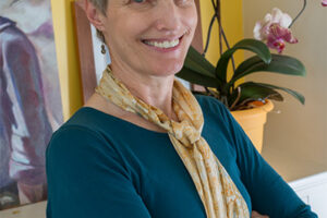 Smiling woman with short gray hair and arms crossed, wearing a scarf in front of a yellow wall and orchids.