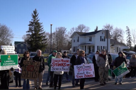 Citizens protest a landfill expansion in Seneca Falls, holding signs like 'Seneca Meadows Stinks' near residential homes.