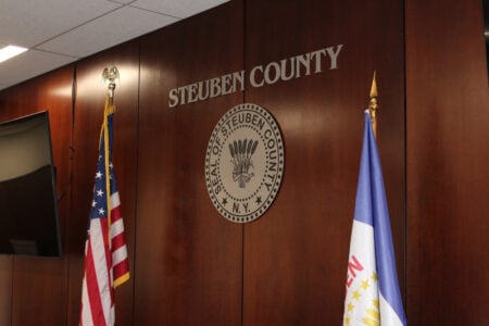 Steuben County seal and name on a wooden wall, flanked by the American flag and another flag.