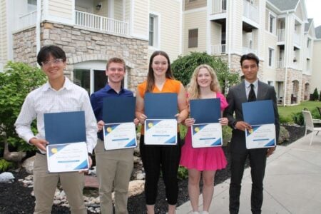 Five smiling scholarship recipients hold certificates outdoors in front of a modern building.