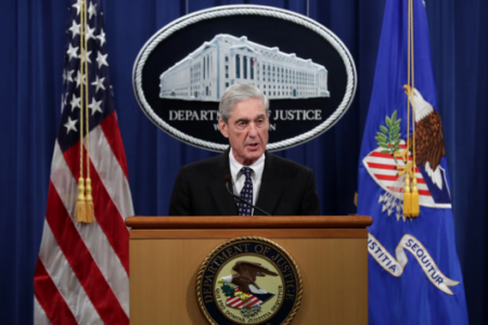 Robert Mueller speaks at a podium with the Department of Justice seal, flanked by US and DOJ flags.