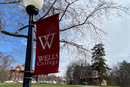 A red Wells College banner with a white 'W' hangs from a lamppost on a tree-lined campus with buildings in the background.