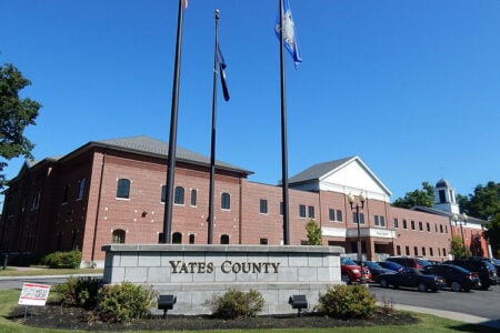Exterior of the Yates County Office Building with flags and a sign on a sunny day.