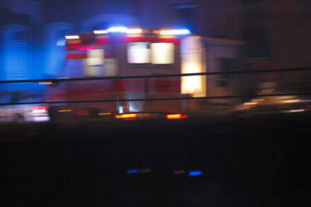 A blurred ambulance with red and blue flashing lights speeds by at night.