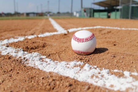 A baseball sits on the brown dirt infield next to a white foul line on a sunny day.