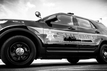 Black-and-white photo of a Cayuga County Sheriff's SUV parked on asphalt under a cloudy sky.