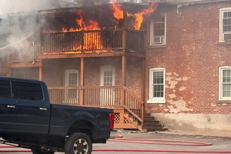 Flames engulf the second-story balconies of a brick building; a dark pickup truck and fire hoses are in the foreground.
