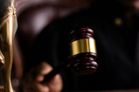 A person's hand holds a wooden gavel with a gold band, with a blurred statue of Lady Justice in the foreground.