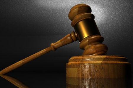 A wooden gavel with a gold band rests on a reflective dark surface, next to its wooden sound block.