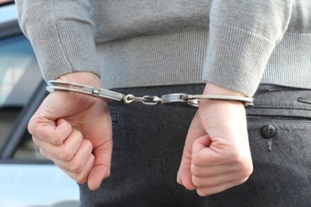 Person in gray sweater with hands cuffed behind their back, next to a vehicle.