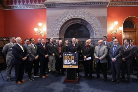 Officials hold a press conference about A.10430 "Better Safety, Better Staffing" in an ornate room.