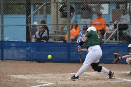 A softball player in a green and white uniform swings at a pitch, with the ball in mid-air and spectators in the background.