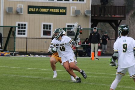 Keuka College player #46 runs with a lacrosse stick on a field, with the Lyle P. Corey Press Box in the background.